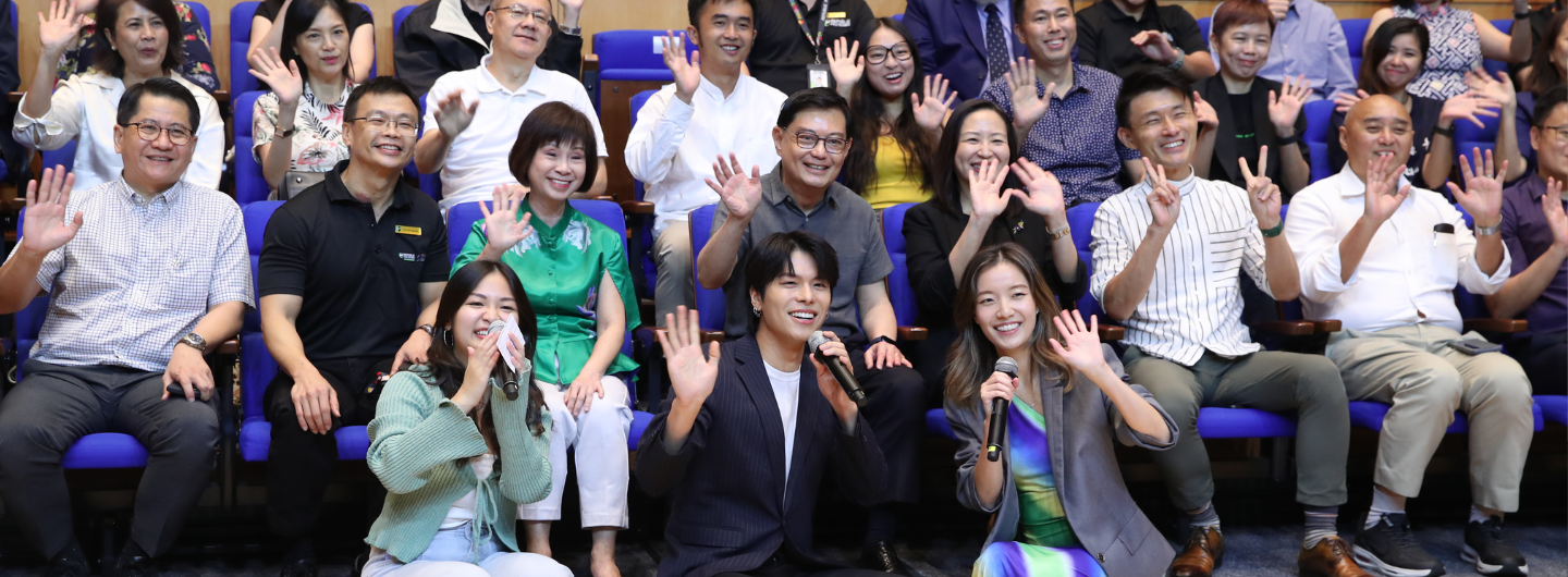 Audience in rows waving at camera, performers in front holding microphones.